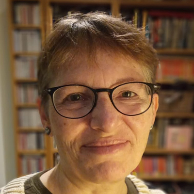Smiling photo of author Marion Wallace, who is wearing glasses and standing in front of a bookcase.